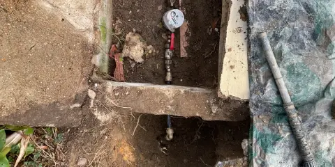 Réparation d'une fuite d'eau : un compteur et des tuyaux sont visibles dans une excavation, avec une bâche de protection à proximité.