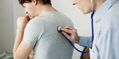 Arzt untersucht Patienten mit Stethoskop. Der Patient hustet, was auf Kurzatmigkeit hindeuten könnte.