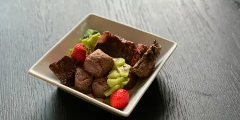 En skål med matberikning för hund: köttbitar, kiwi och jordgubbar. En näringsrik och god måltid för din fyrbenta vän.