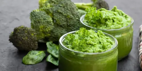 Dwa słoiczki z zielonym puree z brokułów, ozdobione natką pietruszki, na tle świeżych brokułów i liści szpinaku.