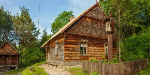 Drewniane chaty i kapliczka na słupie w jednym z **skansenów w Polsce**. W tle las i błękitne niebo.