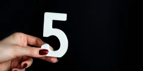 Dłoń z ciemnymi paznokciami trzyma białą cyfrę 5, symbolizującą numerologiczna 5 mężczyzna, na czarnym tle.