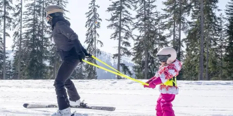 Dziecko w kombinezonie narciarskim uczy się jeździć na nartach, trzymając żółtą linkę.