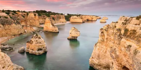 Felsformationen ragen aus dem ruhigen Wasser der **Lagoa, Portugal**, empor. Der Himmel ist in Rosa- und Orangetönen gefärbt, was auf einen wunderschönen Sonnenuntergang hindeutet.