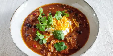 Gęste, aromatyczne chili con carne z fasolą, mięsem i przyprawami, udekorowane śmietaną, startym serem i świeżą kolendrą.