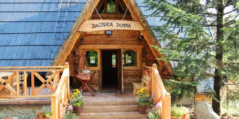 Drewniana bacówka na Jamnej zaprasza do wejścia. Schody prowadzą do otwartych drzwi, a po bokach kwitną kwiaty.