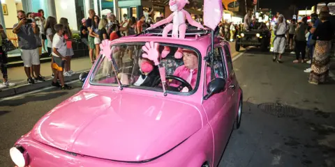 Un'auto rosa shocking sfila a Rimini durante la Notte Rosa, con un pupazzo a forma di pantera rosa sul tetto e un uomo al volante vestito a tema.