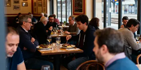 Des clients dînent dans une brasserie de gare, avec des assiettes de viande et des verres de vin sur la table.