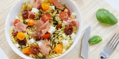 Une salade de pâtes originale avec des tomates cerises, du prosciutto, des dés de mozzarella et une sauce verte savoureuse.