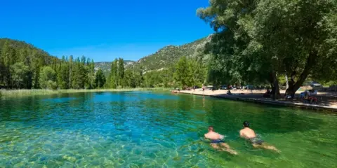 Dos hombres nadan en la cristalina piscina natural Cañamares, rodeada de vegetación y montañas.