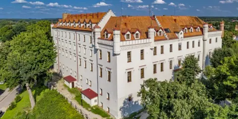 Biały zamek z czerwonym dachem, otoczony zielenią, oferuje wyjątkowe noclegi w zamkach i pałacach. Idealne miejsce na romantyczny wypad.