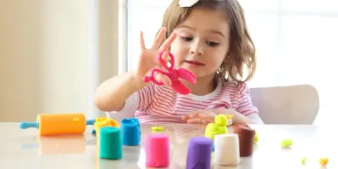 Bambina gioca con pasta modellabile colorata, forbici rosa e mattarello arancione. Giochi di manipolazione Montessori per sviluppare creatività.