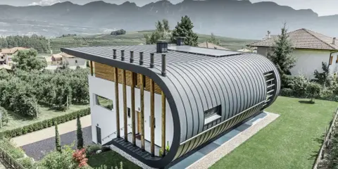 Nowoczesny dom z zaokrąglonym dachem z aluminium, które nie koroduje, otoczony zielenią i górami.