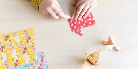 Hände falten Papier für eine Origami Kugel Anleitung. Mehrere bunte Origami-Papierbögen liegen bereit.