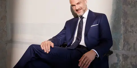 Uśmiechnięty mężczyzna w granatowym garniturze, z krawatem o klasycznej szerokości, siedzi w fotelu.