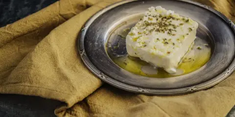 Ein Stück cremiger, vegetarischer Feta mit Kräutern und Olivenöl auf einem silbernen Teller, daneben ein rustikales Leinentuch.