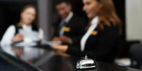 Dzwonek hotelowy na ladzie, za nim recepcjoniści pracują. Wzór karty meldunkowej w hotelu.
