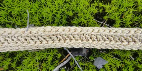 Ett naturfärgat virkat band ligger på grön mossa. Bandet har ett tydligt flätmönster.