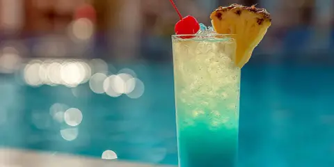 En blå och gul drink med körsbär och ananas står vid en pool. Perfekt för en solig dag.