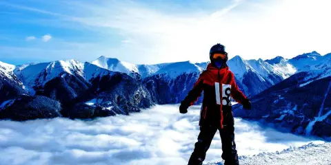 Snowboardzista w czerwonej kurtce softshell i czarnych spodniach hardshell stoi na szczycie góry, nad morzem chmur, z widokiem na ośnieżone szczyty.