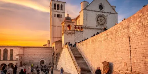 Assisi cosa vedere: la Basilica di San Francesco al tramonto, con la sua imponente facciata e la torre campanaria, illuminata da una calda luce dorata.
