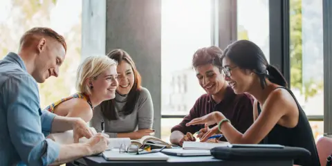 Grupa studentów dyskutuje przy stole, szukając inspiracji, gdzie studiować informatykę. Radość i zaangażowanie w nauce.
