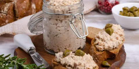 Słoik pełen pysznej pasty z tuńczyka, gotowej do rozsmarowania na chlebie. Na desce kromki z pastą i kaparami, obok gałązka rozmarynu.