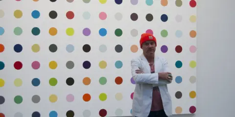 Damien Hirst steht vor seiner berühmten Punkt-Malerei, gekleidet in eine orangefarbene Mütze und einen weißen Blazer.