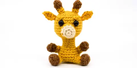 Tierna jirafa amigurumi amarilla, sentada con sus patitas marrones. Sus ojos negros y hocico beige le dan un aspecto adorable.