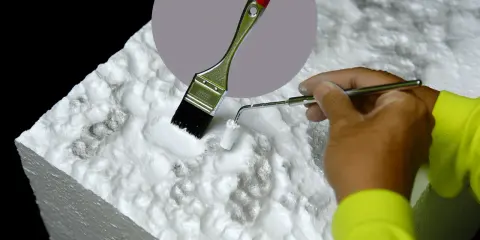 Ręka w żółtej bluzie maluje białą farbą styropian za pomocą pędzla i małego wałka. Dowiedz się, czym pomalować styropian.
