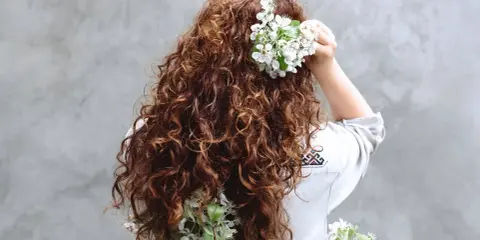 Chevelure brune et bouclée ornée de fleurs blanches, évoquant un décollement de racine naturel et printanier.