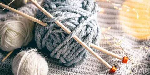 Gruba, szara bukla włóczki z drutami, obok mniejsze kłębki i migoczące światełka.