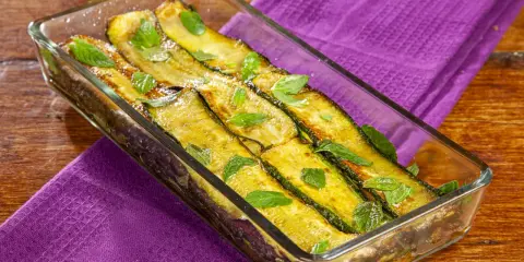 Zucchine alla scapece dorate e profumate alla menta, servite in una pirofila di vetro su un tovagliolo viola.