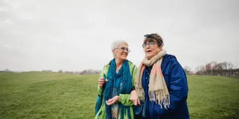Due anziane passeggiano in un prato. Una delle due, con un foulard blu, sembra avere difficoltà a camminare, ma l'altra le offre supporto, un gesto di cura che va oltre l'Alzheimer.