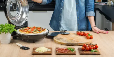 Otwarta opiekacz do pizzy, na kamieniu piec pizza z pomidorkami i bazylią. Obok deska z pomidorkami, startym serem i szynką.
