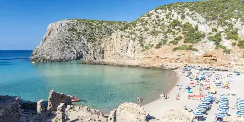 Spiaggia affollata con ombrelloni colorati, acque cristalline e scogliere verdi. Perfetta per vacanze al mare economiche e rilassanti.