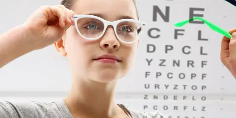 Dziewczynka z wadą wzroku - 20, przymierza okulary na tle tablicy Snellena.