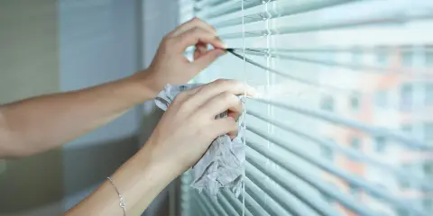 Dłonie z ściereczką wykonują czyszczenie żaluzji. W tle widać rozmyty budynek.