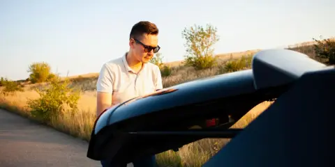 Mężczyzna w okularach przeciwsłonecznych próbuje zrozumieć, jak naprawić zamek w bagażniku, stojąc przy samochodzie na tle pól.
