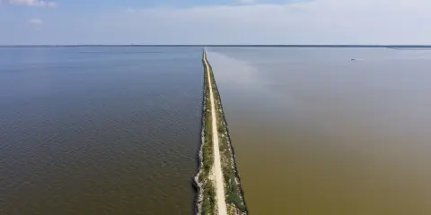 Un sentiero stretto divide due specchi d'acqua, uno scuro e uno più chiaro, sotto un cielo azzurro. Sembra la ciclabile degli angeli.