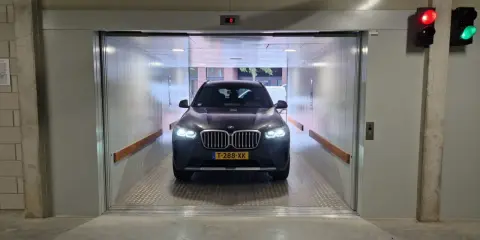 Un coche BMW oscuro espera en un montacoches para garaje. La luz roja del semáforo indica que debe esperar.
