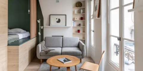 Aménagement petit appartement : lit surélevé, canapé gris, table basse ronde en bois, étagères et grande fenêtre lumineuse.
