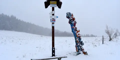 Zimowy krajobraz z ławką i drogowskazem turystycznym. Obok kolorowa rzeźba, być może symbolizująca skopiec szczyt.