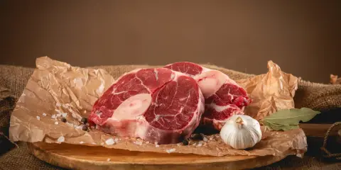 Dwa kawałki surowego ossobuco wołowego, czosnek, liść laurowy i przyprawy na desce.