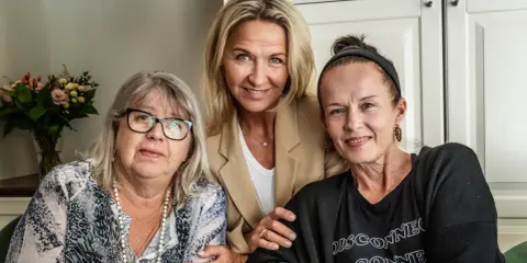 Tre kvinnor, möjligen lill babs barn, ler mot kameran. En kvinna i mitten bär en beige kavaj, den vänstra har glasögon och den högra en svart tröja.