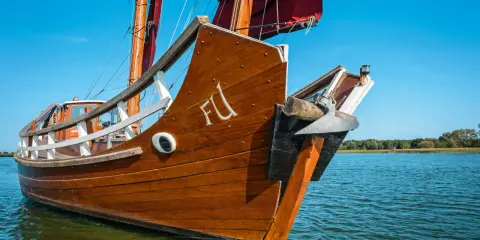 Ein hölzernes Dschunke Schiff gleitet auf ruhigem Wasser unter blauem Himmel. Die Segel sind teilweise gesetzt.