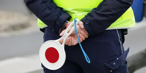 Policjant w kamizelce odblaskowej trzyma za plecami lizak do zatrzymywania pojazdów.