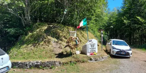Monumento commemorativo con bandiera italiana e corona d'alloro al passo delle radici, circondato da alberi e con un'auto parcheggiata.