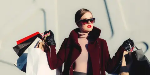 Donna con occhiali da sole e rossetto rosso, avvolta in un cappotto bordeaux, porta borse della spesa, simbolo di moda veloce.