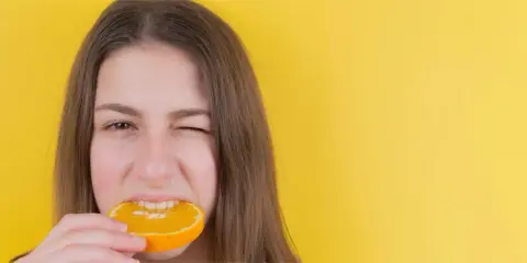 Junge Frau mit einem Stück Orange im Mund, die versucht, nach Weisheitszahn-OP wieder feste Nahrung zu essen.